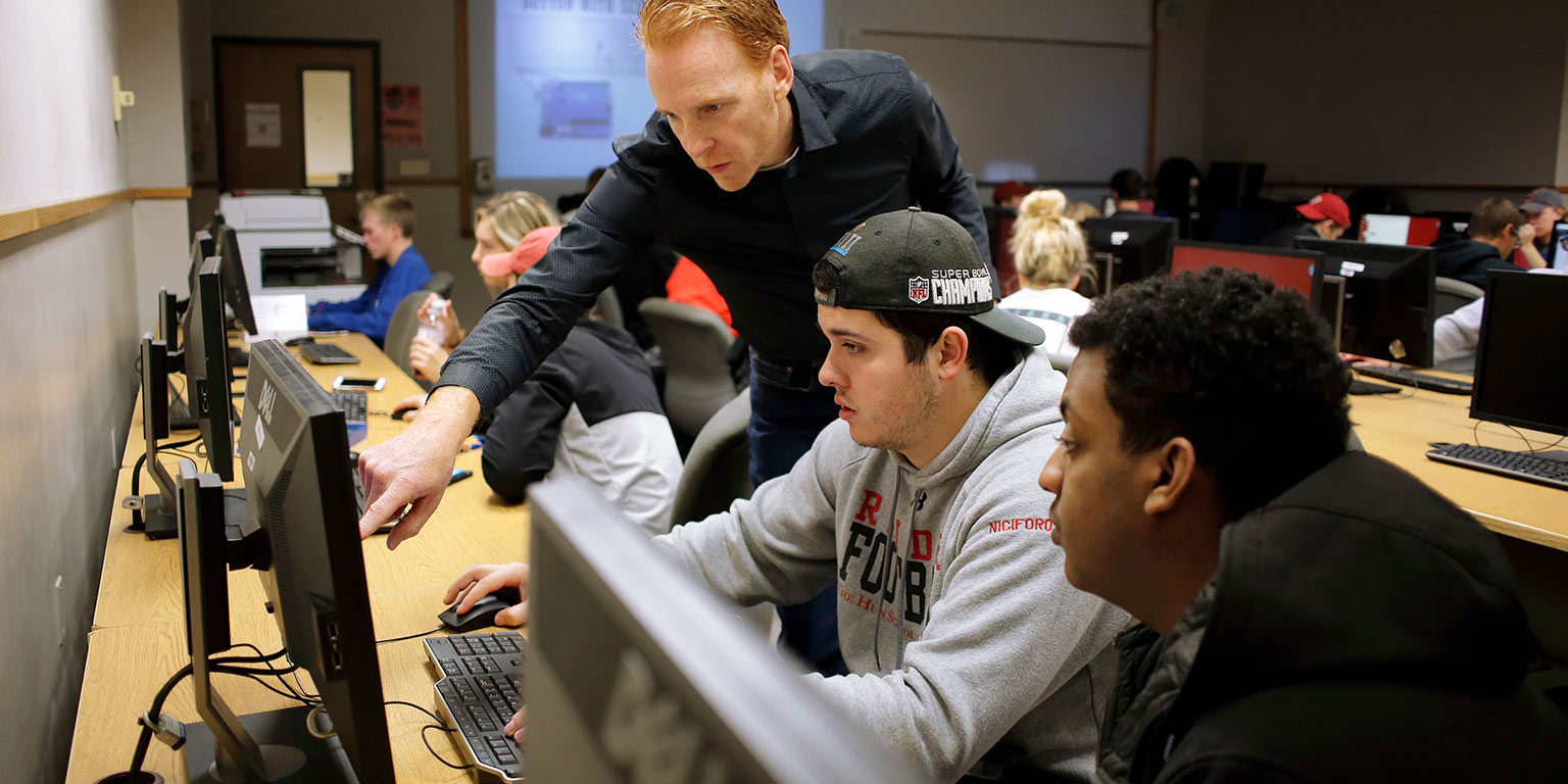 Two students in a computer lab class watch as their professor points to their computer monitor.